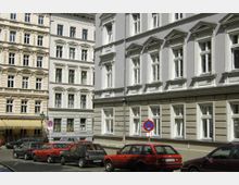 Straßenszene mit historischen Wohngebäuden im Stil des 19. Jahrhunderts, die durch weiße Fensterrahmen und dekorative Fassadenelemente geprägt sind. Im Vordergrund sind geparkte Autos und ein Verkehrsschild mit Parkverbot auf Kopfsteinpflaster zu sehen.