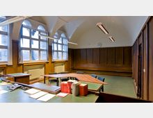 Ein historischer Besprechungsraum mit gewölbter Decke, großen Fenstern mit Sprossen, Holzvertäfelung an den Wänden und einem grünen Tisch mit mehreren Stühlen. Im Vordergrund liegen Bücher und Arbeitsmaterialien auf einem Tisch, während eine an der Wand befestigte Uhr sichtbar ist.