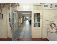 Ein Flur in einem Gebäude, erkennbar durch eine geöffnete doppelflügelige Tür mit Glasfenstern. Der Raum ist gefliest, links und rechts befinden sich Arbeitsflächen und Wanddekorationen; weiter hinten führen Fliesen und Beleuchtung den Blick zu einem helleren Bereich.