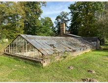 Ein altes Gewächshaus mit verrostetem Metallrahmen und teilweise beschädigten Glasflächen, umgeben von einer grasbewachsenen Wiese und hohen Bäumen im Hintergrund. Das Gebäude hat eine Ziegelmauerbasis und einen gemauerten Schornstein an der Rückseite.