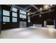 Großer, leerer Innenraum mit schwarzem Industriestil, großen Glasbausteinfenstern, hellbeigem Boden und Metallträgern an der Decke für Beleuchtung. Im Hintergrund befinden sich Glastüren und ein Durchgang zu einem angrenzenden Raum.