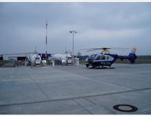 Ein Hubschrauber der Polizei steht auf einer betonierten Fläche neben mehreren Treibstofftanks und Zapfsäulen. Im Hintergrund sind ein flaches Gebäude und ein offenes Feld sichtbar, der Himmel ist bewölkt.