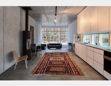 Modernes Wohnzimmer mit offenem Grundriss, gekennzeichnet durch Sichtbetonwände, eine schlichte Küche mit weißen Schränken auf der rechten Seite und einem großen, gemusterten Teppich in der Mitte. Ein Kamin sowie Sitzmöbel stehen vor einer großen Glasfront mit Blick auf einen Wald und einen Außenbereich mit Holzstapel und Hängematte.