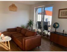 Wohnzimmer mit einem braunen Ledersofa, einem niedrigen weißen Couchtisch und einer Kommode mit einem Globus und Dekorationsgegenständen. Große Glas-Schiebetüren führen zu einem Balkon mit Outdoor-Möbeln, und mehrere Zimmerpflanzen stehen an der Fensterfront. Eine Korblampe hängt von der Decke, und an der Wand hängt ein gerahmtes Bild.