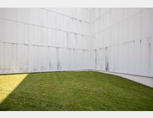 Rasenfläche umgeben von hohen, weißen Wänden aus Betonplatten mit sichtbaren Wasserflecken und einer leichten Kurve am Bodenrand. Der Bereich ist teilweise von Sonnenlicht beschienen.