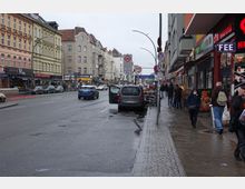 Städtische Straßenszene in einem Einkaufsviertel mit mehrstöckigen Gebäuden, teils mit historischen Fassaden. Entlang der nassen Straße sind geparkte Autos, bunte Geschäfte, Restaurants und Personen auf dem Bürgersteig zu sehen.
