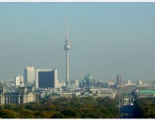 Blick auf das Stadtzentrum von Berlin mit dem Fernsehturm als markantem Wahrzeichen in der Mitte, umgeben von modernen und historischen Gebäuden wie dem Berliner Dom. Im Vordergrund ist der Tiergarten mit dichtem Laub zu sehen, rechts das Brandenburger Tor.