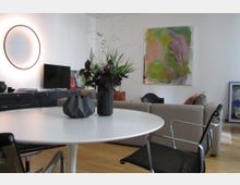 Wohnzimmer mit modernem Design, bestehend aus einem runden weißen Tisch mit einer Vase und Pflanzen darauf, grauem Sofa im Hintergrund, Designerstuhl und Kunstwerken an den Wänden. Ein schwarzer Schrank mit Fernseher und ein beleuchteter runder Spiegel zieren die weiße Wand.
