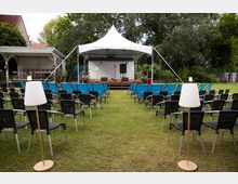 Open-Air-Veranstaltungsbereich auf einer Rasenfläche mit Reihen von Stühlen, die auf eine kleine Bühne unter einem weißen Pavillonzelt ausgerichtet sind. Hinter der Bühne stehen Bäume und Grünflächen, daneben ein überdachter Holzpavillon und ein Gebäude am Rand.