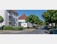 Straßenszene in einem Wohngebiet mit weiß verputzten Mehrfamilienhäusern, roten Ziegeldächern und Balkonen. Die Straße ist von Bäumen gesäumt, mit parkenden Autos auf einer Seite; Straßenschilder und Gehwege sind ebenfalls sichtbar.