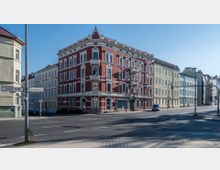 Ein mehrstöckiges Wohngebäude im Stil des späten 19. Jahrhunderts mit rot verputzter Fassade und weißen Stuckelementen an einer Straßenecke. Umgeben von weiteren historischen Gebäuden, verläuft eine breite Straße mit Verkehrszeichen durch die Szene.
