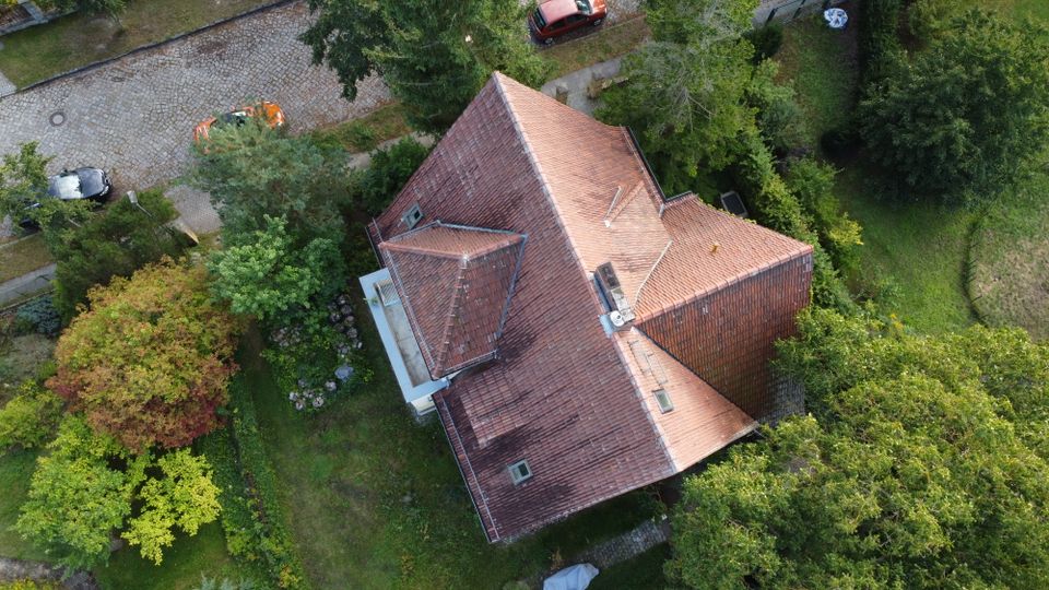 Luftaufnahme eines Hauses mit rotem Ziegeldach und mehreren Dachflächen, umgeben von dichtem Grün und Gartenflächen. Am oberen Bildrand verläuft eine Kopfsteinpflasterstraße mit parkenden Autos entlang eines Gehwegs.