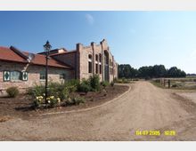 Backsteinbau mit großen Rundbogenfenstern, flankiert von einem kleineren Gebäudeteil mit grünen Fensterläden. Ein geschwungener, unbefestigter Weg führt daran vorbei, im Vordergrund Blumenbeete, im Hintergrund Felder und Bäume.