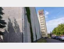 Industriegebäude mit einer hohen, beigefarbenen Turmuhr im Hintergrund, dessen Fassade durch Fensterreihen strukturiert ist. Im Vordergrund eine seitliche, helle Wand mit Bewuchs, daneben ein schmaler Weg mit parkenden Autos und Bäumen.