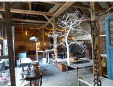 Geräumiger Innenraum einer rustikalen Scheune mit sichtbaren Holzbalken, schrägem Dach und Steinwänden. Der Raum ist mit Holzmöbeln, Vintage-Dekor, einem weiß gestrichenen künstlichen Baum und Lichterketten dekoriert.