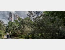 Ein Hochhaus mit moderner Architektur ragt am Horizont zwischen einer weitläufigen grünen Parklandschaft mit Nadelbäumen und dichten Sträuchern empor. Im Vordergrund spaziert ein älteres Paar auf einem schmalen Weg durch den Park.