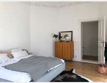 Schlafzimmer in einer Wohnung mit hellen Wänden, einem Doppelbett mit weißer Bettwäsche und grauer Decke, Holzboden und einer Kommode aus Holz. Auf der Kommode stehen eine Lampe, eine Pflanze und ein gerahmtes Bild; daneben ist eine geöffnete weiße Tür sichtbar.