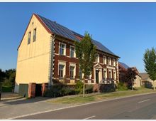Zweigeschossiges Gebäude im klassischen Stil mit gelber und roter Klinkerfassade, großen Fenstern und einem Satteldach mit Solarzellen, gelegen an einer Straße mit Bäumen und Gehweg. Im Vordergrund eine kleine Treppe und ein gepflegter Garten.