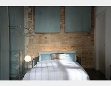 Minimalistisch eingerichtetes Schlafzimmer mit einer freistehenden Backsteinwand, einem Doppelbett mit weißen und blauen Bettdecken sowie einem grauen Kopfteil. Links steht ein Metallregal mit einer Hängelampe und einem Kleiderbügelständer.