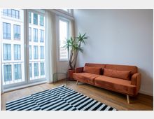 Wohnzimmer mit Holzboden, einem orangefarbenen Sofa, einem schwarz-weiß gestreiften Teppich und einem Glas-Beistelltisch. Große Fenster mit weißen Vorhängen bieten Blick auf ein modernes Gebäude gegenüber.