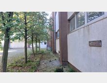 Außenbereich eines Gebäudes mit beiger und brauner Fassade, samt rechteckigen Fenstern. Auf der linken Seite stehen mehrere Bäume auf einem gepflasterten und teilweise bewachsenen Weg; im Hintergrund ist ein Hof mit weiteren Gebäudeteilen sichtbar.