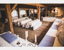 Rustikaler Innenraum eines Veranstaltungsraums mit Holzbalken und Ziegelboden. Weiße, mit Tischdecken bedeckte Bänke und Sitzpolster sind um einen zentralen Bereich angeordnet, dekoriert mit Blumen und Kerzen.