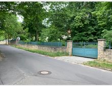 Eine asphaltierte Straße führt an einer Steinmauer entlang, hinter der hohe Bäume stehen. Die Mauer ist mit grün gestrichenen Metallzäunen und einem großen Tor aus Metall, flankiert von steinernen Pfeilern, versehen.