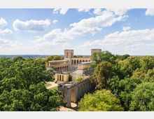 Historisches Gebäude mit neoklassizistischer Architektur, bestehend aus einer mehrschichtigen Terrassenanlage mit Arkaden und Türmen, eingebettet in eine grüne Parklandschaft unter einem klaren Himmel mit einigen Wolken. Im Hintergrund ist eine weite, leicht hügelige Landschaft zu sehen.