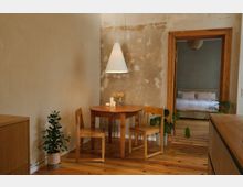 Gemütlicher Wohnbereich mit einem kleinen runden Holztisch und zwei Holzstühlen, beleuchtet von einer weißen Hängelampe. Im Hintergrund befindet sich eine offene Tür, die in ein Schlafzimmer mit einem weißen Bett führt; links und rechts sind Zimmerpflanzen zu sehen.