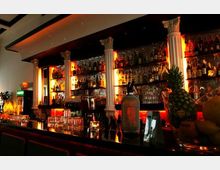 Eine Bar mit dunkler Theke, beleuchtetem Regal voller Flaschen und gläsernen Getränken in warmem, orangefarbenem Licht. Die Einrichtung umfasst weiße Säulen und Dekorationen mit tropischen Früchten wie Ananas und Zitronen.