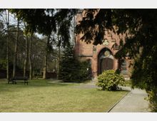 Backstein-Kapelle in einem Park, umgeben von Bäumen und Sträuchern. Vor der Kapelle führt ein gepflasterter Weg, daneben eine Bank und eine offene Rasenfläche.