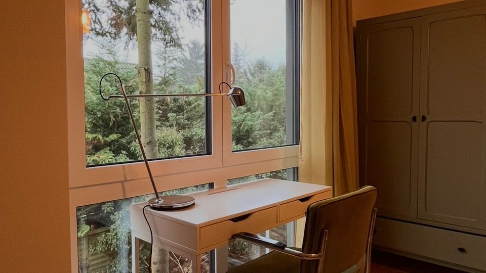 Ein kleiner Arbeitsbereich mit einem weißen Schreibtisch und einem Stuhl mit Metallrahmen, vor einer großen Glasfront mit Blick auf einen Garten mit Bäumen. Rechts im Raum steht ein großer, grauer Schrank, und der Boden ist mit Holzdielen ausgelegt.