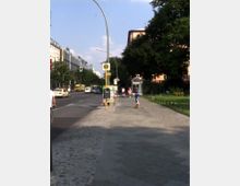 Städtische Straße mit Gehweg und Fahrradweg, flankiert von grünen Bäumen und mehrstöckigen Gebäuden. Im Vordergrund befindet sich eine Bushaltestelle mit einem Haltestellenschild, im Hintergrund sind Autos und Fußgänger zu sehen.