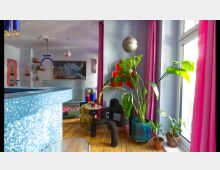 Innenraum eines modern und künstlerisch gestalteten Raums mit bunten Möbeln und Dekoration. Zu den sichtbaren Elementen gehören eine Sitzgruppe, große Zimmerpflanzen in Töpfen, pinke Vorhänge, ein glitzernder Tresen und eine Discokugel an der Decke.