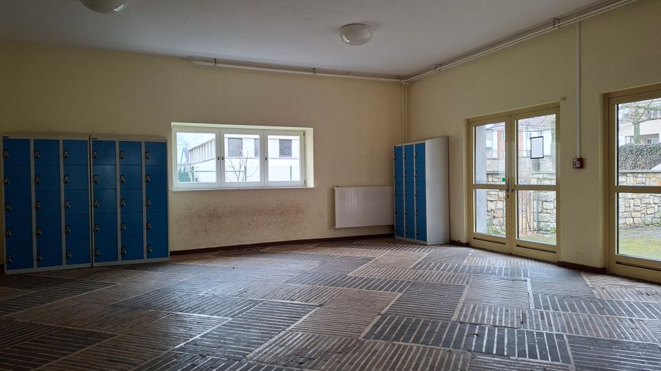 Leerer Umkleide- oder Garderobenraum mit beigen Wänden, blau-grauen Schließfächern an den Seiten und einem Boden aus dunklen Ziegelplatten im Muster. An einer Wand befindet sich ein Fenster, daneben eine Glastür nach außen mit Blick auf eine Steinmauer.