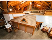 Innenansicht einer großen Scheune oder Lagerhalle mit hohem Holzdachstuhl und einer umlaufenden Galerie im Obergeschoss. Unten steht ein kleiner Holzbau mit Satteldach und Dreiecksfenster, umgeben von Betonboden, gelagerten Möbeln und Arbeitsmaterialien.