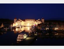 Hafenanlage bei Nacht mit beleuchtetem Gebäude im Hintergrund, das wie ein großes Hotel oder Resort wirkt, umgeben von kleineren Ferienhäusern am Wasser. Im Vordergrund liegen Boote am Steg, und die Szene spiegelt sich im ruhigen Wasser wider.