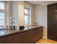 Moderne Küche mit dunklen Holzunterschränken, einer Arbeitsplatte aus braunem Marmor und einer kleinen Spüle. Zwei Fenster lassen Tageslicht herein, daneben stehen Küchengeräte wie ein Toaster und Wasserkocher, während eine Holzlattenwand den Raum abtrennt.