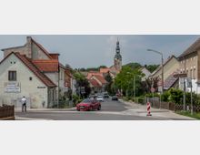 Straßenszene in einer Kleinstadt mit Einfamilienhäusern, einem roten Auto im Vordergrund und einem Kirchturm mit Spitzdach im Hintergrund. Verkehrsschilder und Gehwege säumen die Straße, die von Bäumen und gepflegten Vorgärten umgeben ist.