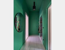 Schmaler Flur mit intensiv grünen Wänden und weißem Holzboden, einem runden Wandspiegel links, einer schwarzen Deckenlampe und Türen zu benachbarten Räumen, darunter ein Raum mit floraler Tapete.