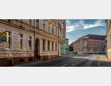 Straßenszene in einer kleinen Stadt mit historischen Gebäuden. Links ein pastellfarbenes Gebäude mit Verzierungen und Schildern, rechts ein Eckgebäude mit orangefarbener Fassade und roten Dachziegeln; die Straße ist leer und von Gehwegen gesäumt.