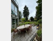 Rückseite eines Wohnhauses mit einer gepflasterten Terrasse und einem großen ovalen Tisch, umgeben von mehreren Stühlen. Im Hintergrund befindet sich ein Garten mit Rasen, Bäumen, Pflanzen und Sitzgelegenheiten, darunter eine Holzbank und ein kleiner Tisch.