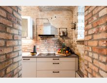 Kleine Küche mit Sichtziegelwänden, weißem Unterschrank und dunkler Arbeitsplatte. An der Wand befinden sich ein eingebauter Dunstabzug, ein Regal mit Gewürzen sowie eine Obstschale mit frischen Früchten auf der Arbeitsfläche.