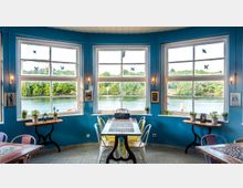 Ein heller Innenraum eines Cafés mit blauen Wänden, großen Fenstern und Blick auf einen See mit grünen Bäumen. Vor den Fenstern stehen kleine Tische mit Stühlen und dekorativen Pflanzen.