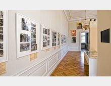 Ein heller Museumsraum mit Holzboden und klassischer Wandvertäfelung. An den Wänden hängen gerahmte Schwarz-Weiß-Fotografien sowie bunte Poster, und rechts befindet sich eine gläserne Vitrine.