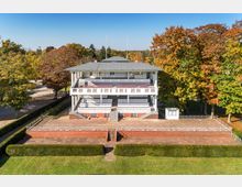 Historisches Tribünengebäude aus weißem Holz mit drei offenen Ebenen, umgeben von Herbstbäumen. Davor liegen gepflasterte Terrassen mit niederer Begrenzung und gepflegtem Rasen.