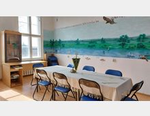 Kleiner Seminarraum mit einem langen Tisch, umgeben von blauen Stühlen. An der Wand befindet sich eine großflächige Wandmalerei mit einer Flusslandschaft, Bäumen und Tieren, dazu ein dekorativer Schriftzug. Im Raum stehen ein Holzschreibtisch und ein Glaskabinett.