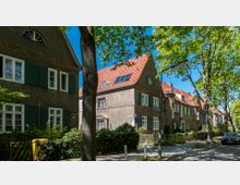 Straßenzug in einem Wohngebiet mit historischen Reihenhäusern, die braune Fassaden, rote Ziegeldächer und weiße Fenster mit grünen Fensterläden haben. Im Vordergrund stehen Bäume, die Schatten auf den Gehweg werfen, und einzelne Elemente wie ein Straßenschild und ein parkendes Auto sind sichtbar.
