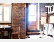 Gemütliche Küche mit rustikalem Stil: Wände aus Ziegelmauerwerk, Holztür mit Fenster, ein Holzstuhl und ein kleiner Tisch stehen links. Rechts befindet sich ein weißer Herd und eine Hängeschrankschublade. Tageslicht fällt durch die Fenster und die Tür.