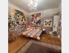 Schlafzimmer mit einer markanten, künstlerischen Gestaltung: eine Wand mit rauer, teilweise abblätternder Oberfläche, teilweise bedeckt mit großen, bunten Gemälden und Collagen. Ein Doppelbett mit einem gemusterten Bettbezug steht auf einem Holzboden, der teilweise mit einem Orientteppich bedeckt ist. Ein Kronleuchter hängt von der Decke.